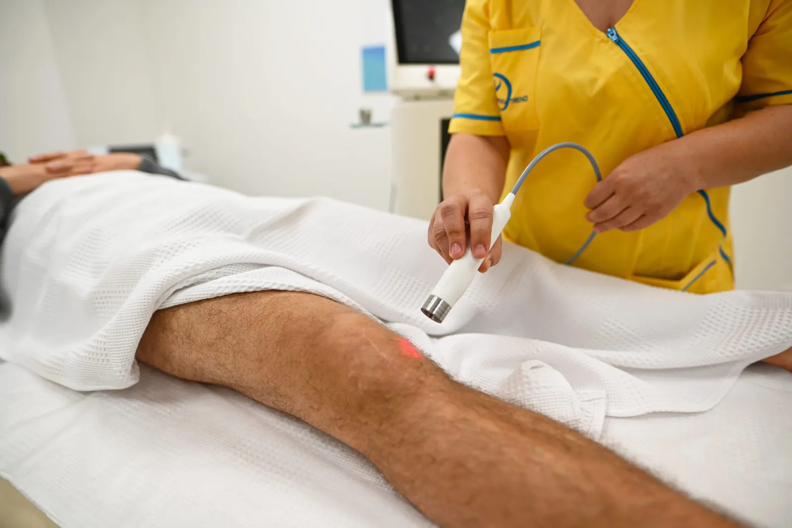 Terapeuta kézi lézerfejjel kezeli a páciens térdét a kezelőágyon